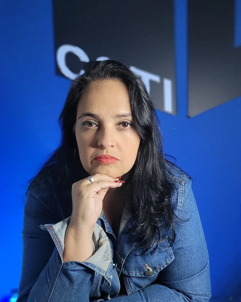 Fernanda Nunes em frente ao logo da COTI Informática, com jaqueta jeans e a mão apoiada no queixo