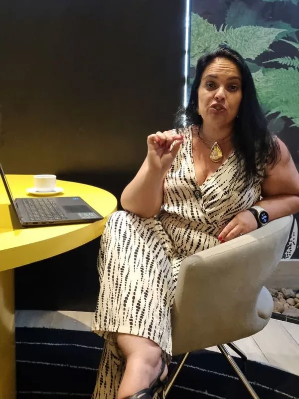 Fernanda Nunes sentada ao lado de uma mesa com notebook, explicando algo com um gesto de mão